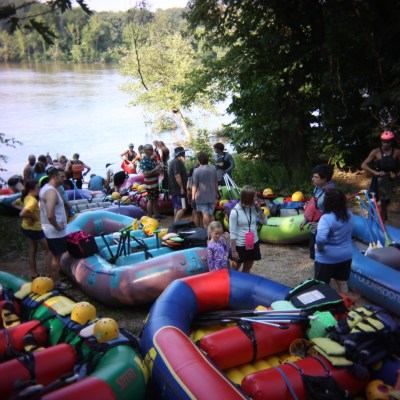 Group of people preparing for rafting by the river, surrounded by colorful inflatables and equipment.