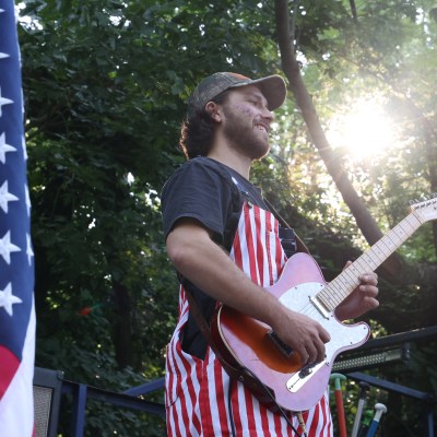 Guitarist in star-striped outfit plays beside American flag and trees with sunlight filtering through.