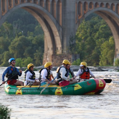 Group of people rafting on a river near arched bridge, wearing helmets and life jackets.