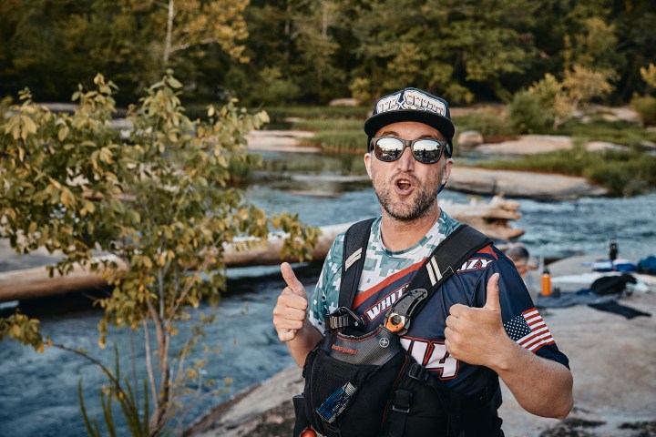 Man in sunglasses and cap gives thumbs up near river with trees in background.