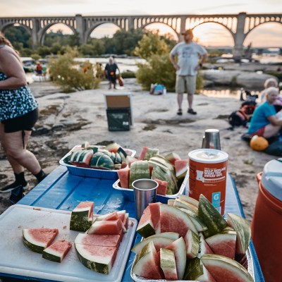 Slices of watermelon on a picnic table at sunset near a bridge with people in the background.