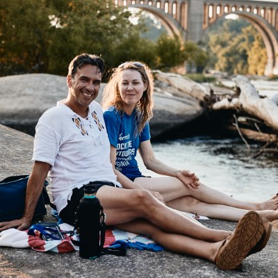 Two people sitting on a rock near a river, with a bridge in the background.
