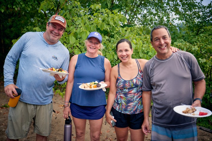 Four people smiling, standing outdoors and holding plates with food.