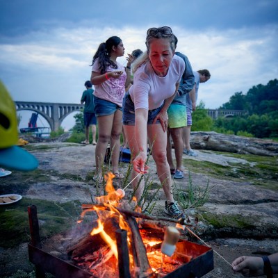 People roasting marshmallows over a fire pit with a bridge in the background.