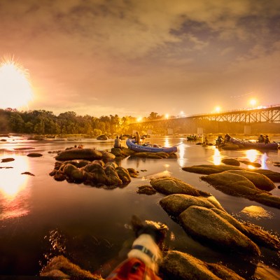 People in rafts on a river under a bridge with fireworks in the sky at sunset.