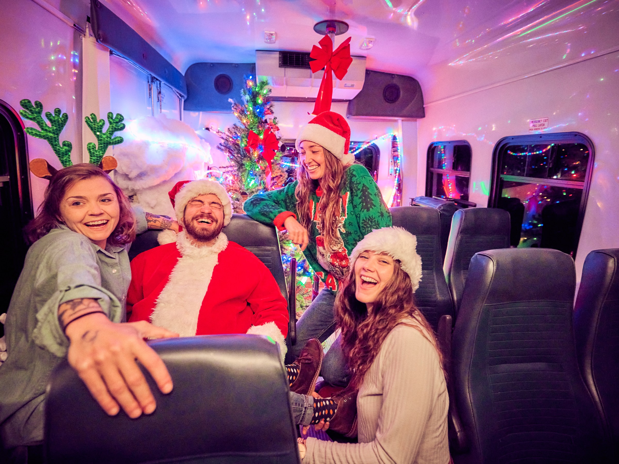 Four people in festive attire laughing inside a decorated bus with Christmas lights and tree.