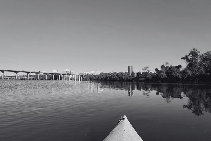 Black and white view from a kayak on a calm river, with a distant bridge and city skyline.